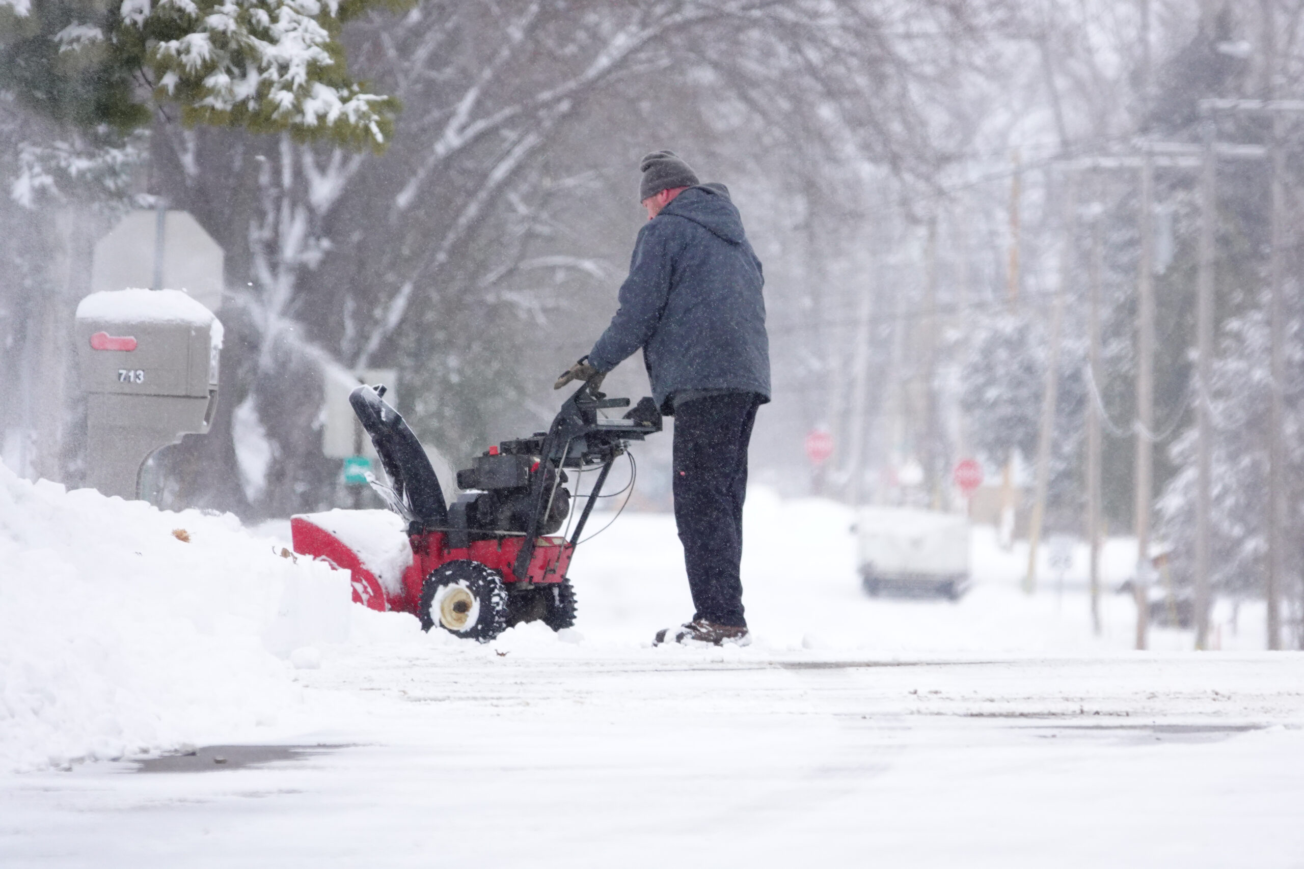 A middle-aged man in a blue-grey hoodie and black pants using a red snow blower to clear a pile of snow off of a road.<br />
