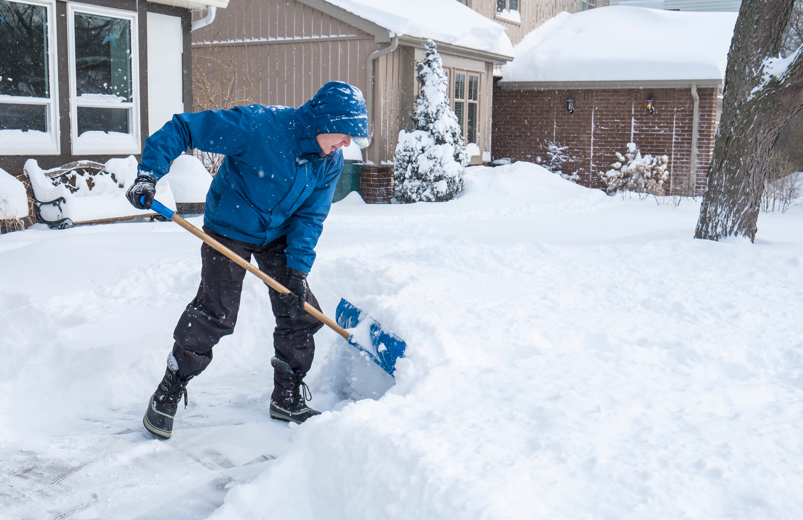 A man in a blue jacket Tinley Park IL performing a snow removal with a blue shovel in a snowy landscap background with a front yard of a residential home in the background.<br />
