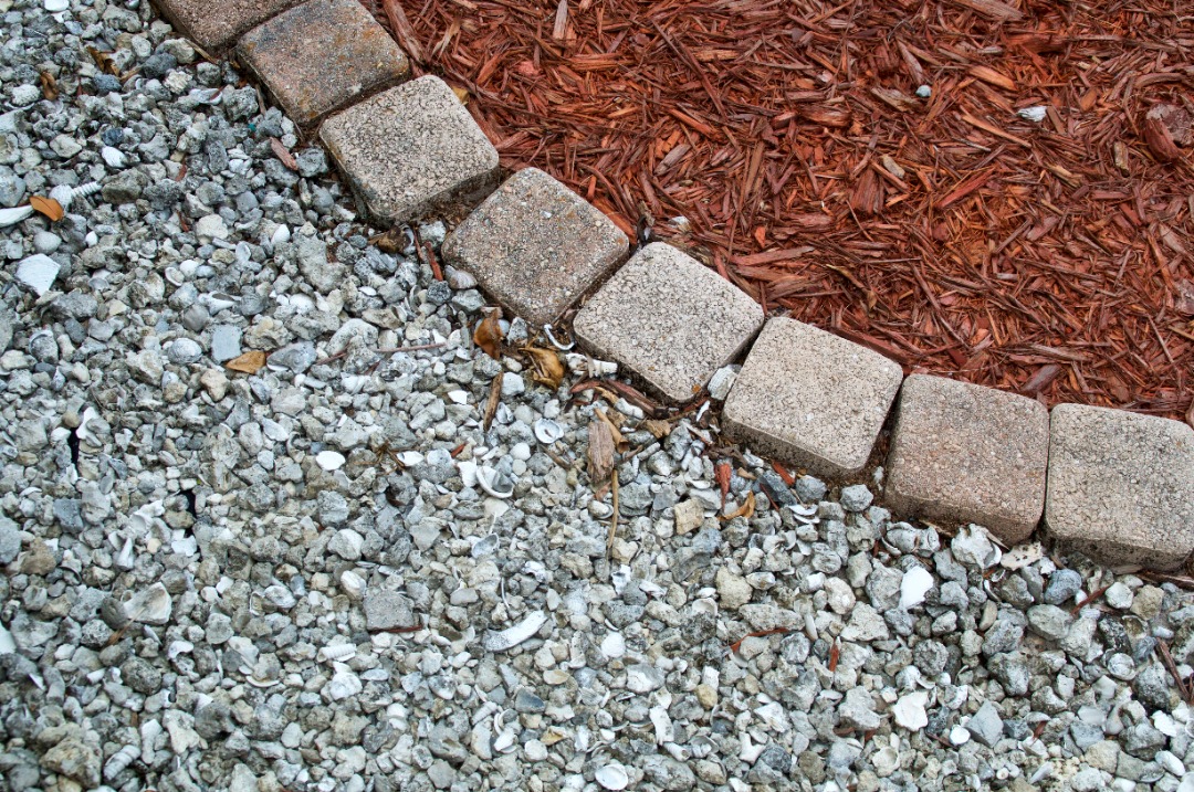 Close-up of landscaping materials including gray gravel, red mulch, and beige paving stones forming a decorative border.