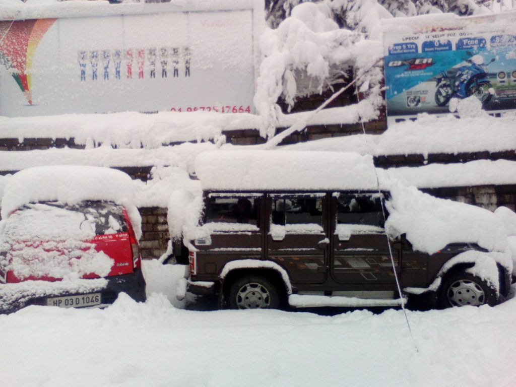 Two snow-covered vehicles, a red hatchback and a dark SUV, parked on a snowy street with thick snow on their roofs and windshields. A snow-laden wall and billboards are visible in the background.